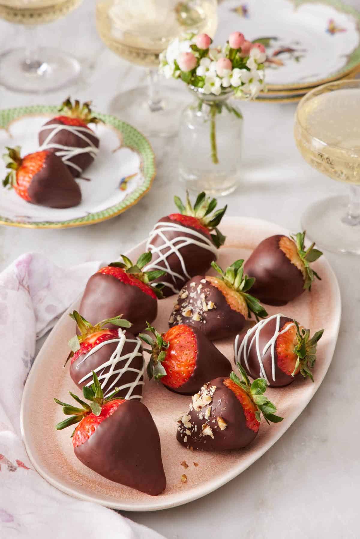 A platter of chocolate covered strawberries with a small vase of flowers and small plate with two chocolate covered strawberries in the background.
