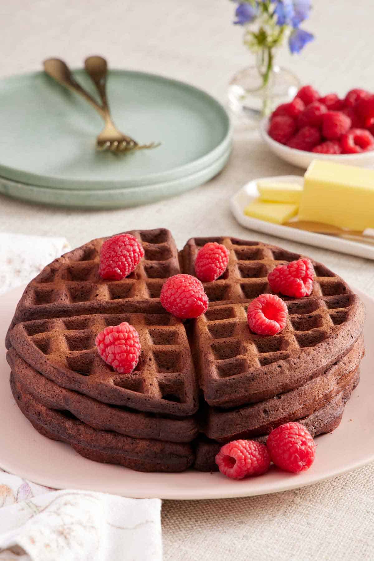 A stack of three chocolate waffles topped with fresh raspberries. A stack of plates, butter, and raspberries in the background.