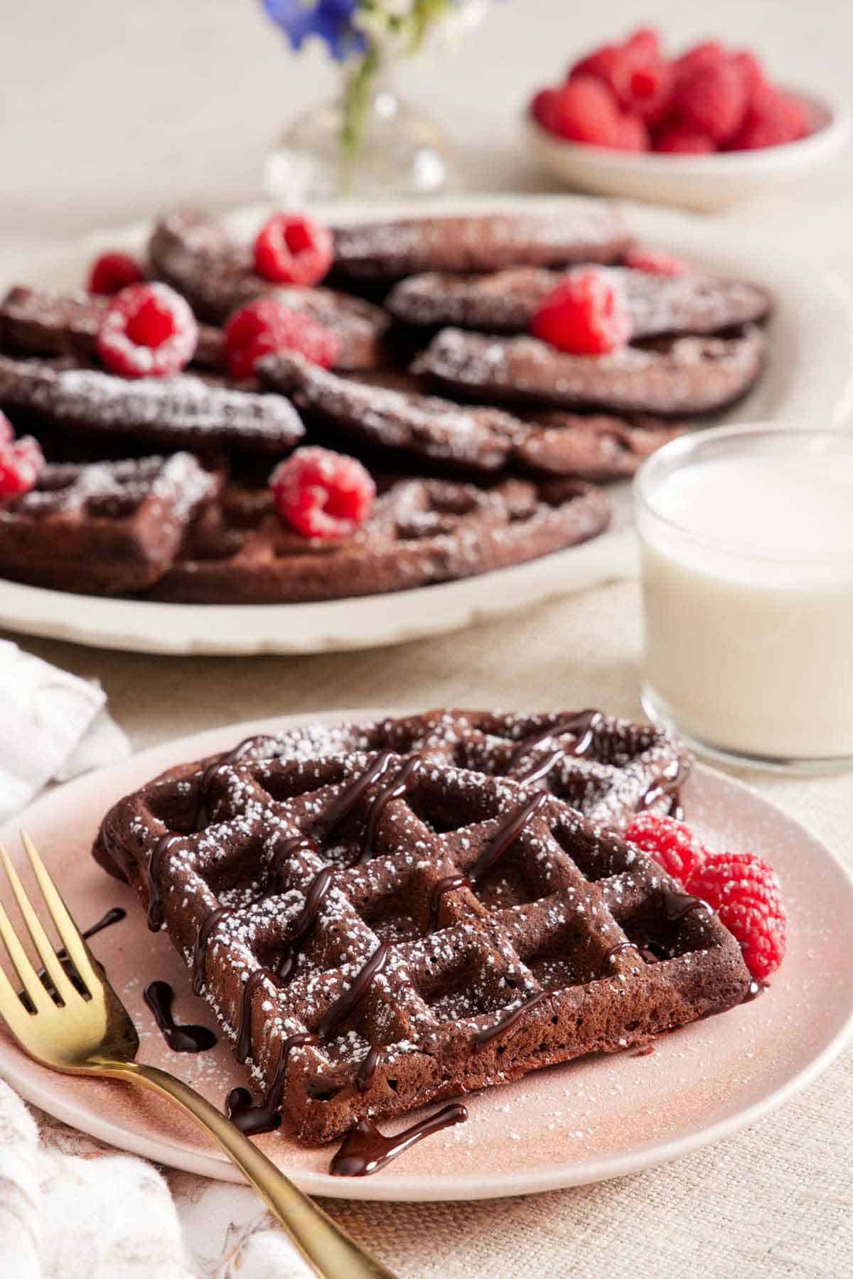 A plate with a piece of chocolate waffles topped with chocolate and powdered sugar with a fork on the side. A glass of milk and platter of more chocolate waffles in the background.