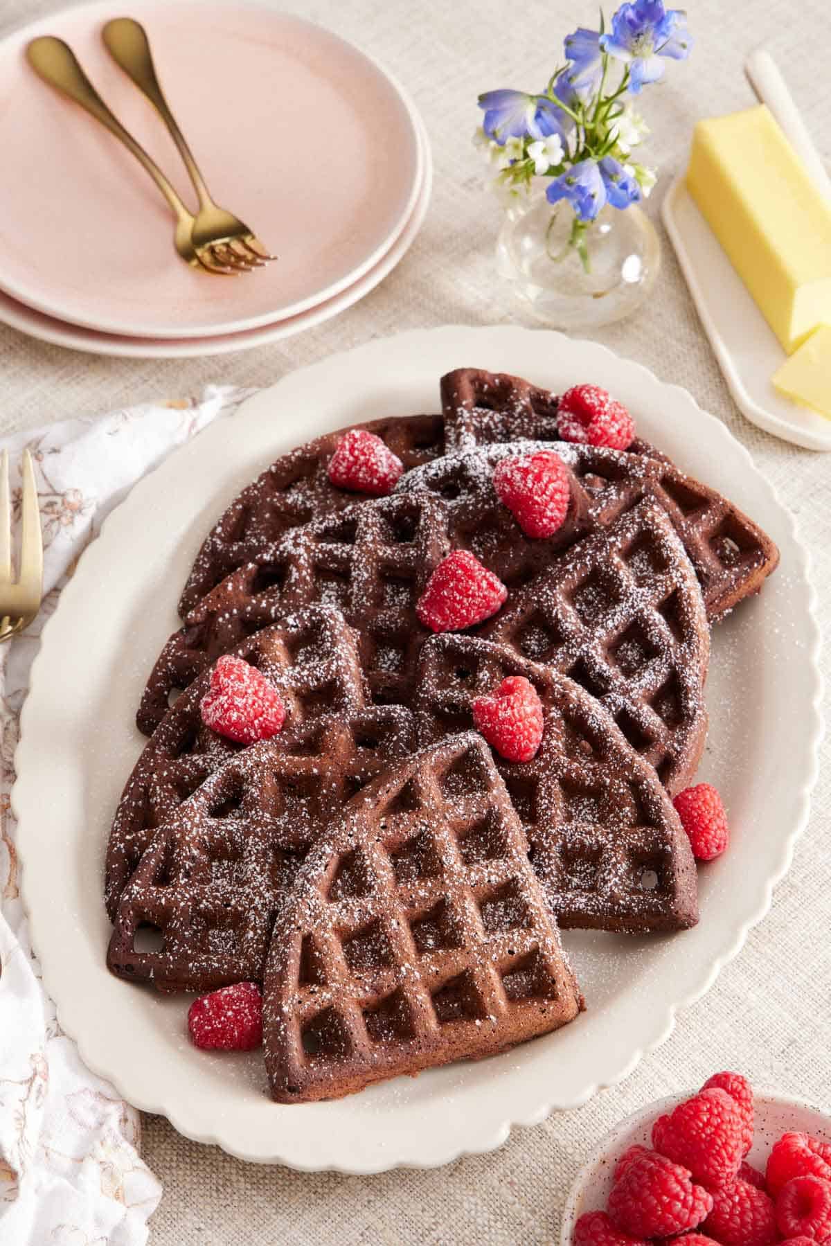 An overhead view of a platter of chocolate waffles topped with powdered sugar and raspberries. More raspberries, butter, flowers, plates, and a fork scattered around.