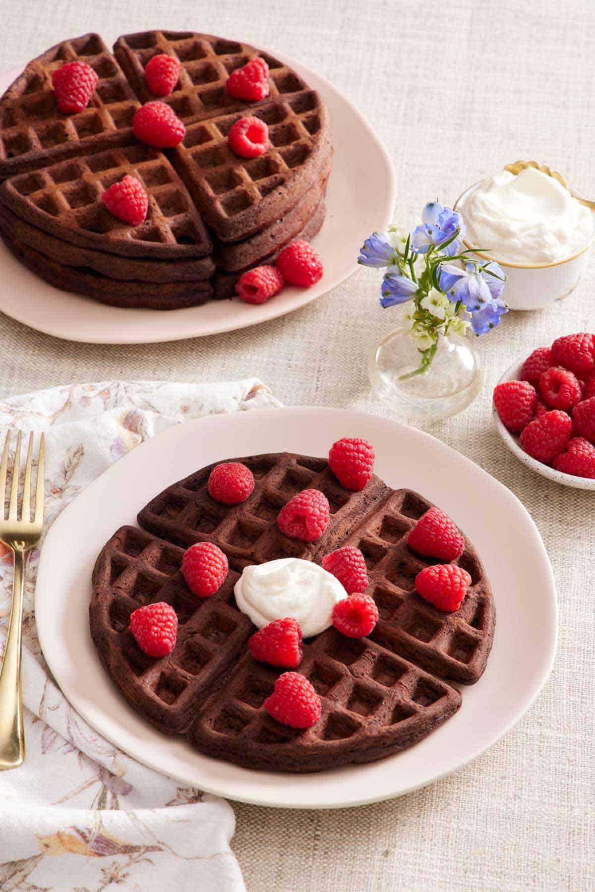 A plate with a chocolate waffle topped with some whipped cream and fresh raspberries. A stack of more waffles topped with raspberries, a bowl of raspberries, a vase of flowers, and a bowl of whipped cream in the back.