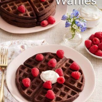 Pinterest graphic of a plate with a chocolate waffle topped with some whipped cream and fresh raspberries. A stack of more waffles topped with raspberries, a bowl of raspberries, a vase of flowers, and a bowl of whipped cream in the back.