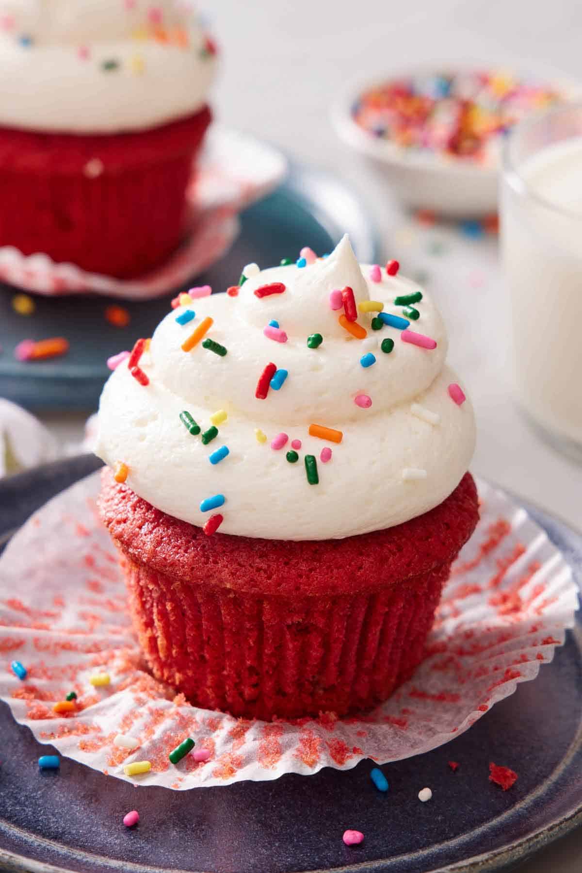 A red velvet cupcake with the paper liner pulled down and with ermine frosting topped with rainbow sprinkles. A glass of milk, bowl of sprinkles, and another cupcake in the background.