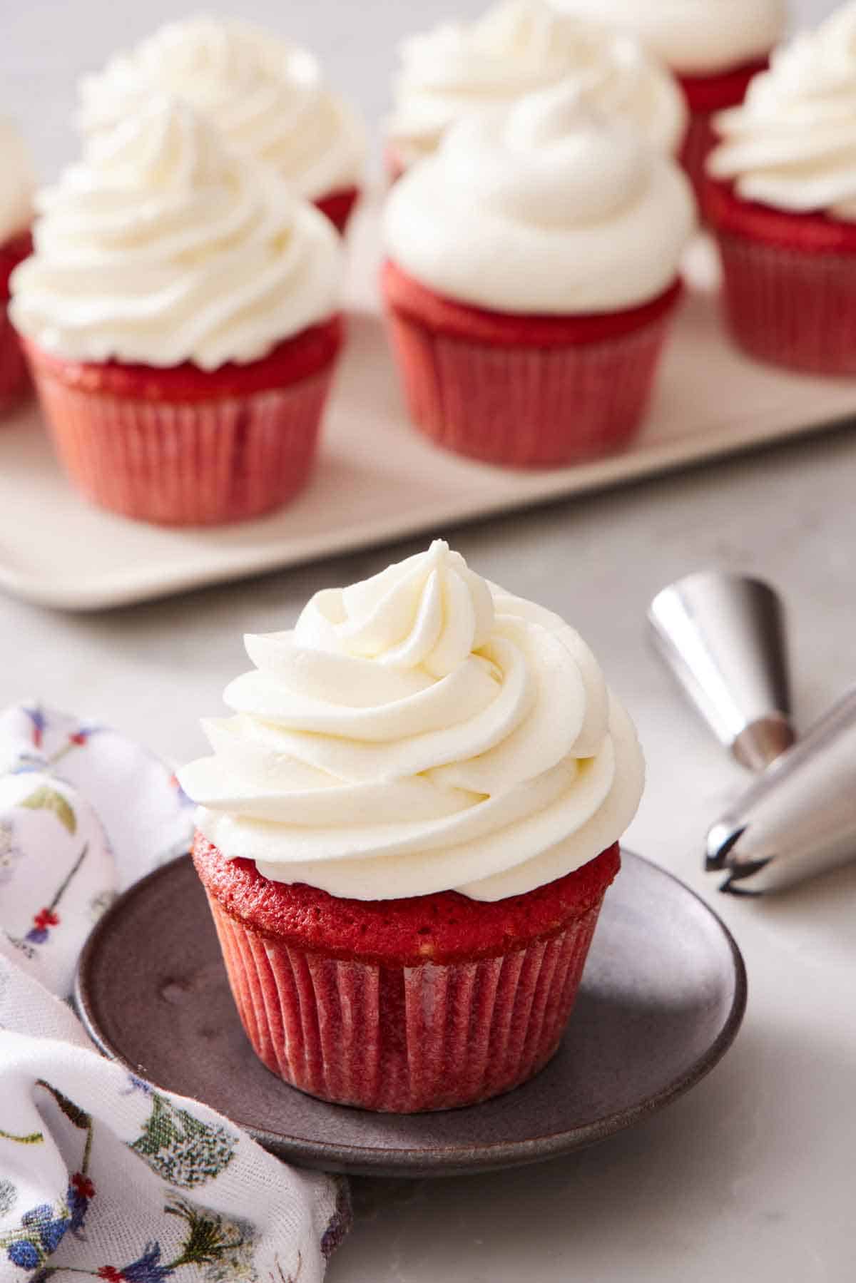 A red velvet cupcake with ermine frosting. More frosted cupcakes in the background and piping tips.