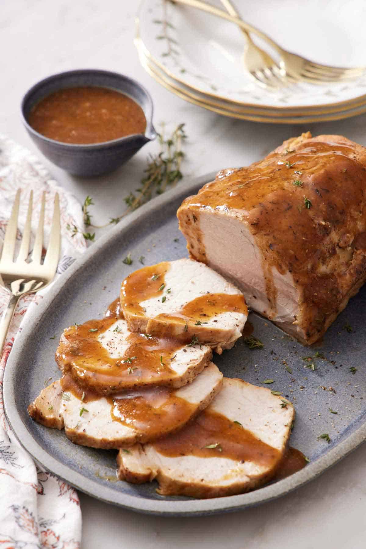 A platter of Instant Pot pork loin with four pieces sliced in front with sauce drizzled on top. A bowl of gravy, stack of plates, and forks in the background.