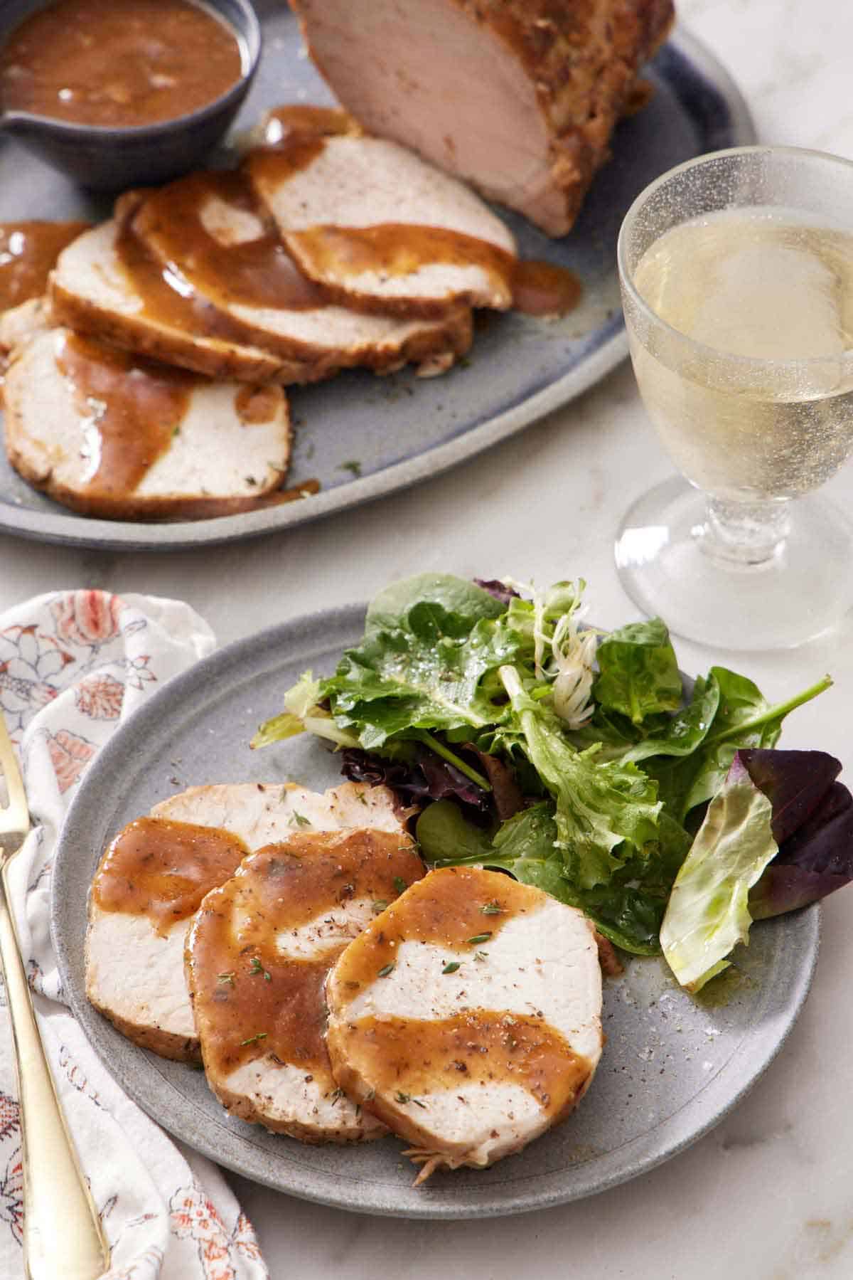 A plate with three slices of Instant Pot pork loin with sauce and salad. A drink in the back along with more sliced pork.