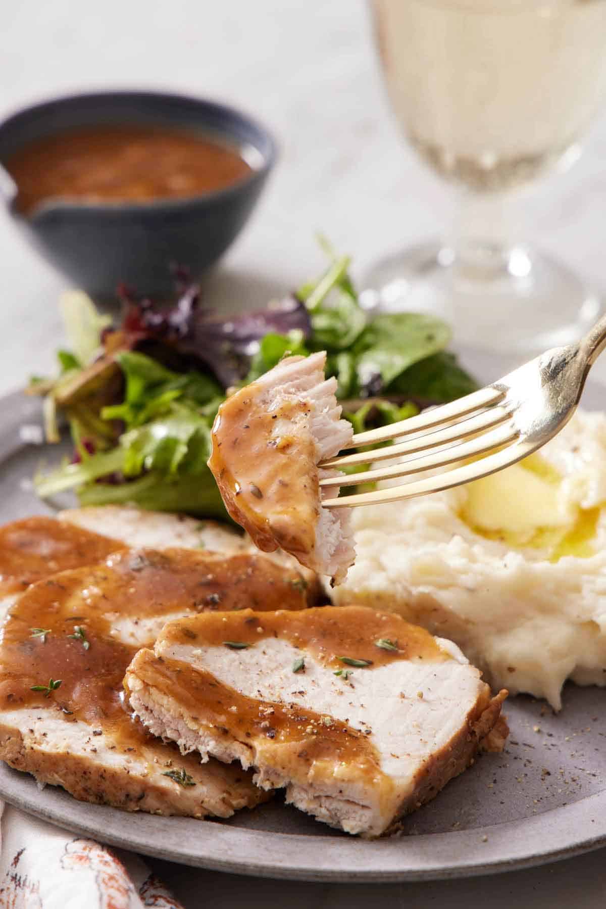 A fork lifting up a bite of Instant Pot pork loin from a plate with pork, mashed potatoes, and salad. Gravy drizzled on top and a bowl with more in the background.