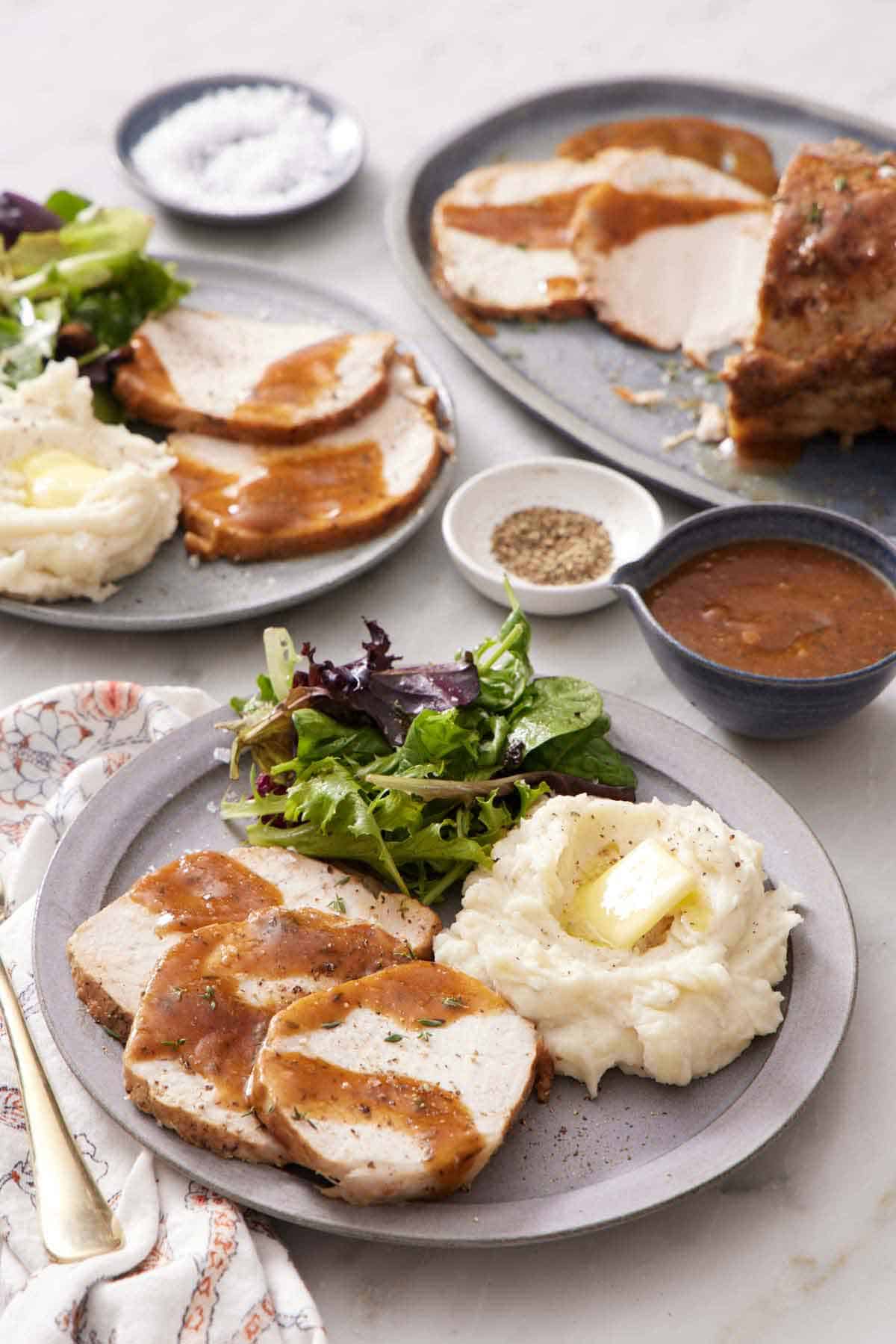 A plate with a serving of Instant Pot pork loin with gravy, mashed potatoes, and salad. Another plated serving in the back along with the rest of the pork, a bowl of gravy, salt, and pepper.