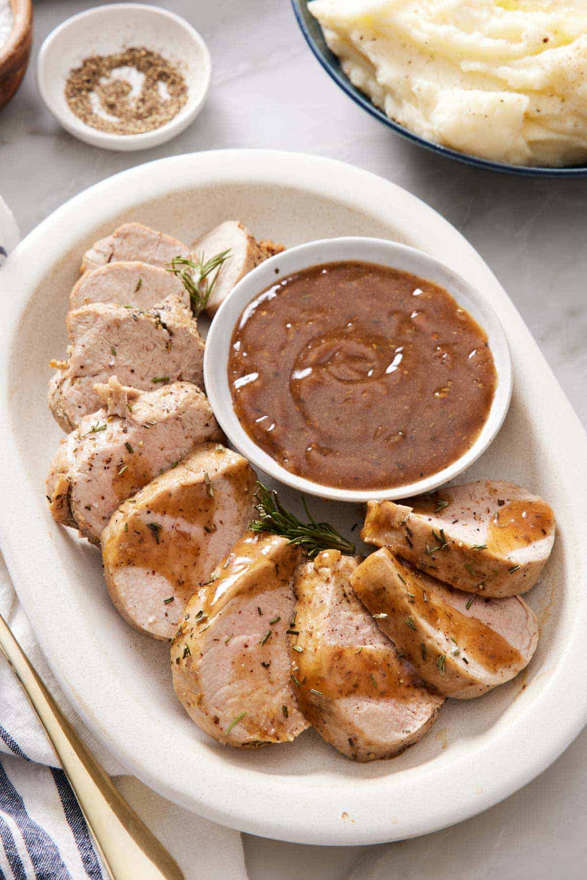 Overhead view of a platter of sliced Instant Pot pork tenderloin with a bowl of gravy. A bowl of pepper in the back along with mashed potatoes.