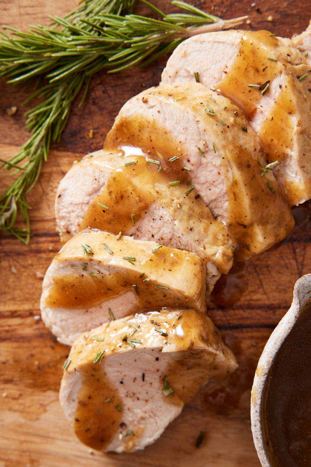 A sliced Instant Pot pork tenderloin on a wooden serving board with gravy drizzled on top with rosemary on the side.