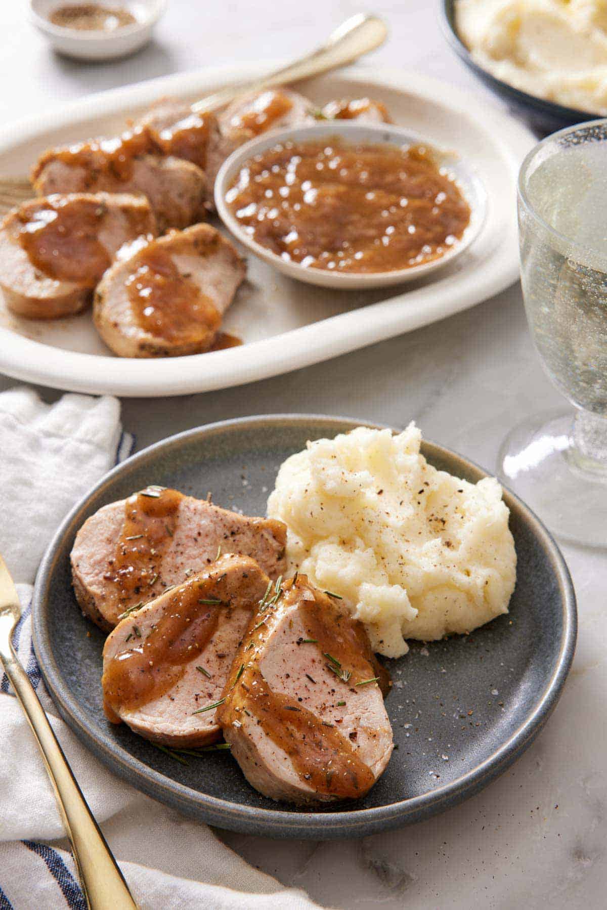 A plate with three pieces of Instant Pot pork tenderloin on a plate with mashed potatoes. A platter with more sliced tenderloin in the background along with a bowl of gravy.
