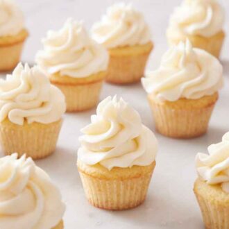 Multiple mini cupcakes with vanilla frosting on a marble surface with a plate of sprinkles on the side.