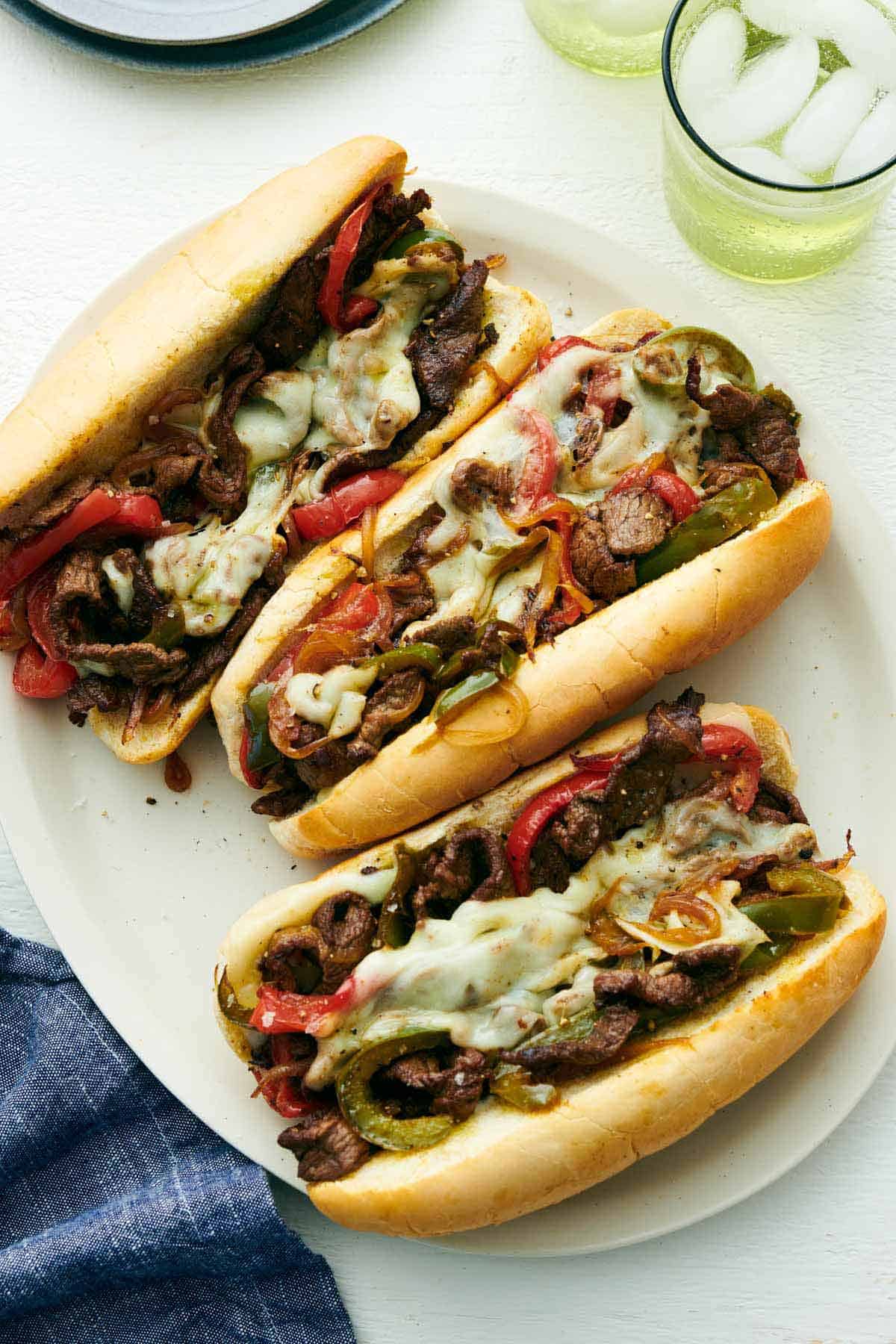 Overhead view of three Philly cheesesteak sandwiches on a platter. A drink off to the side.