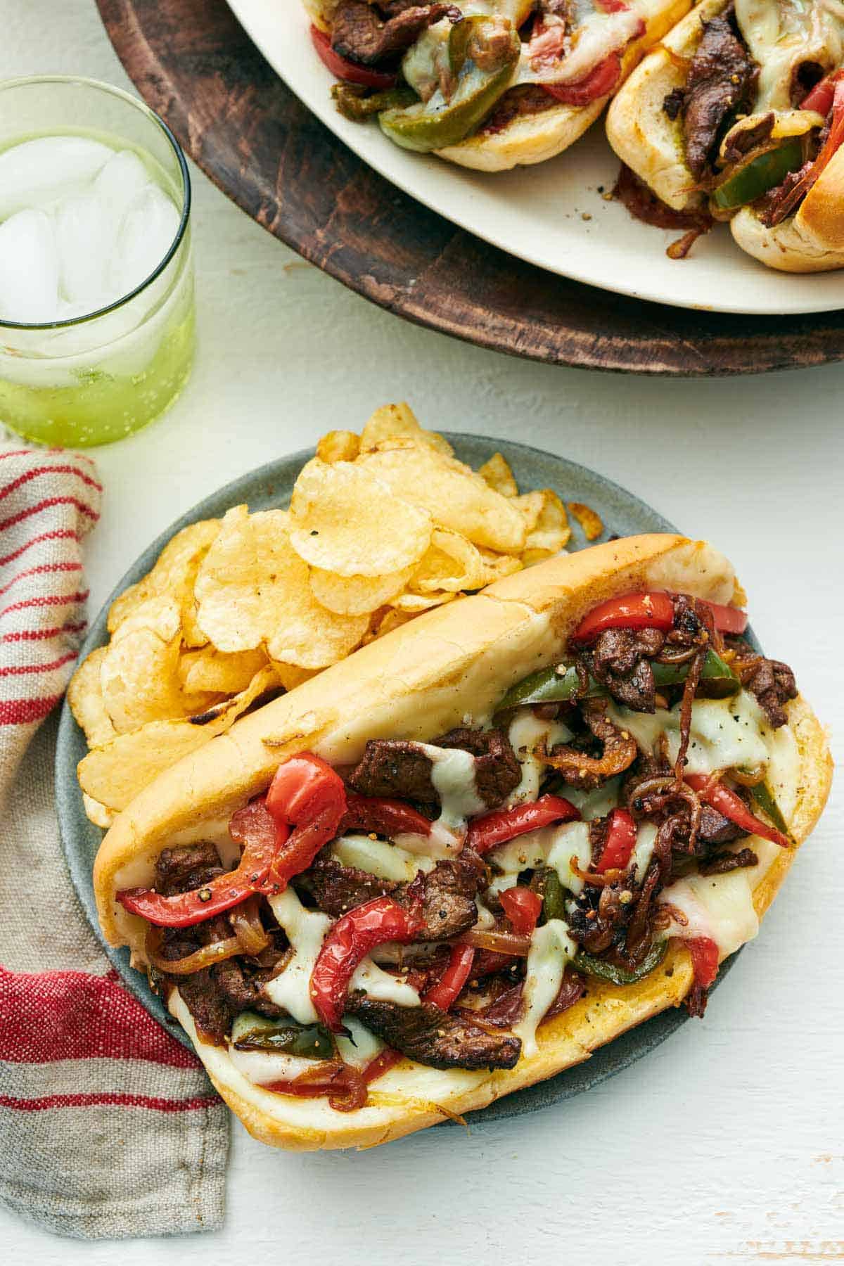 A plate of Philly cheesesteak with chips. A drink in the back and more sandwiches off to the side.