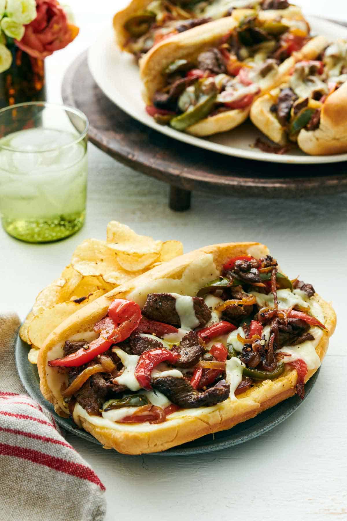 A Philly cheesesteak on a plate along with chips. A drink in the back along with a platter of three more Philly cheesesteaks.
