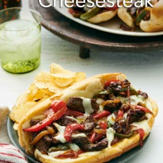 Pinterest graphic of a Philly cheesesteak on a plate along with chips. A drink in the back along with a platter of more Philly cheesesteaks.