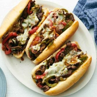 Overhead view of a platter with three Philly cheesesteaks. A plate and napkin on the side.
