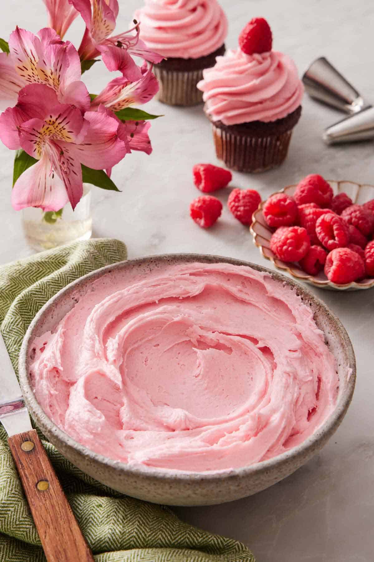 A bowl of raspberry buttercream. Pink lilies, fresh raspberries, and frosted cupcakes in the background.