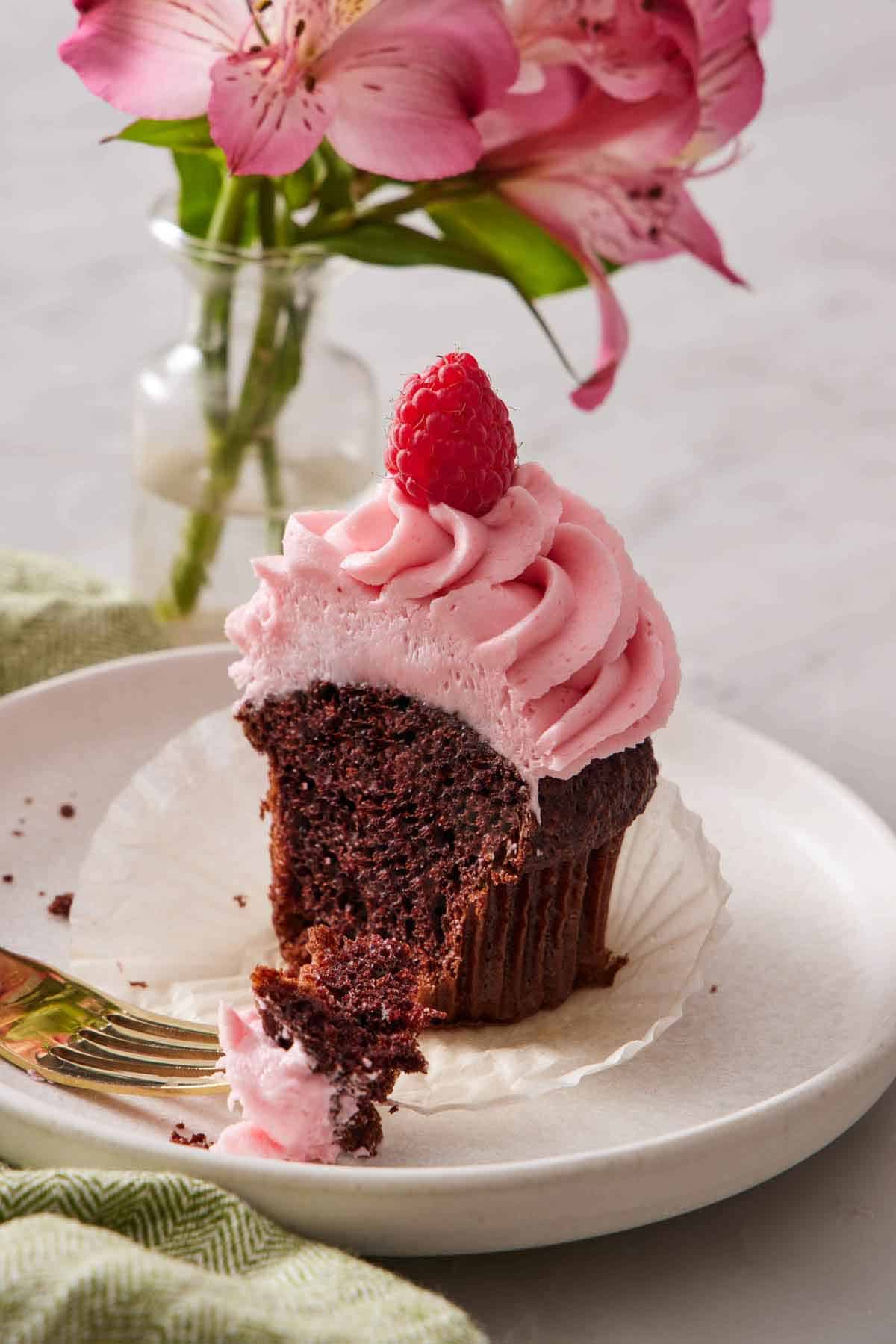 A plate with a cupcake topped with raspberry buttercream and a fresh raspberry with a bite taken out.
