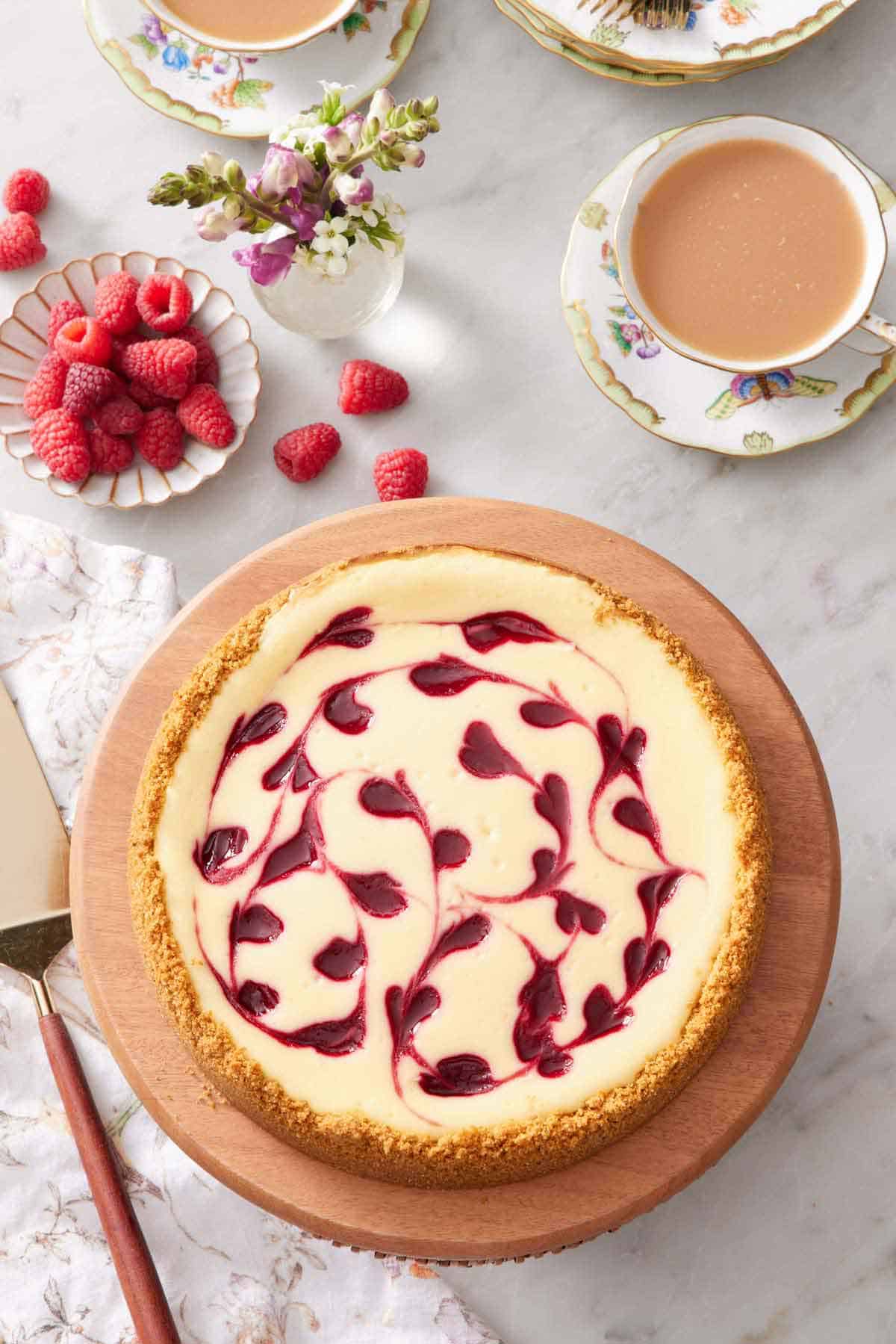 Overhead view of a raspberry cheesecake on a wooden cake stand. A plate of fresh raspberries, vase of flowers, and two cups of milk tea off to the side.