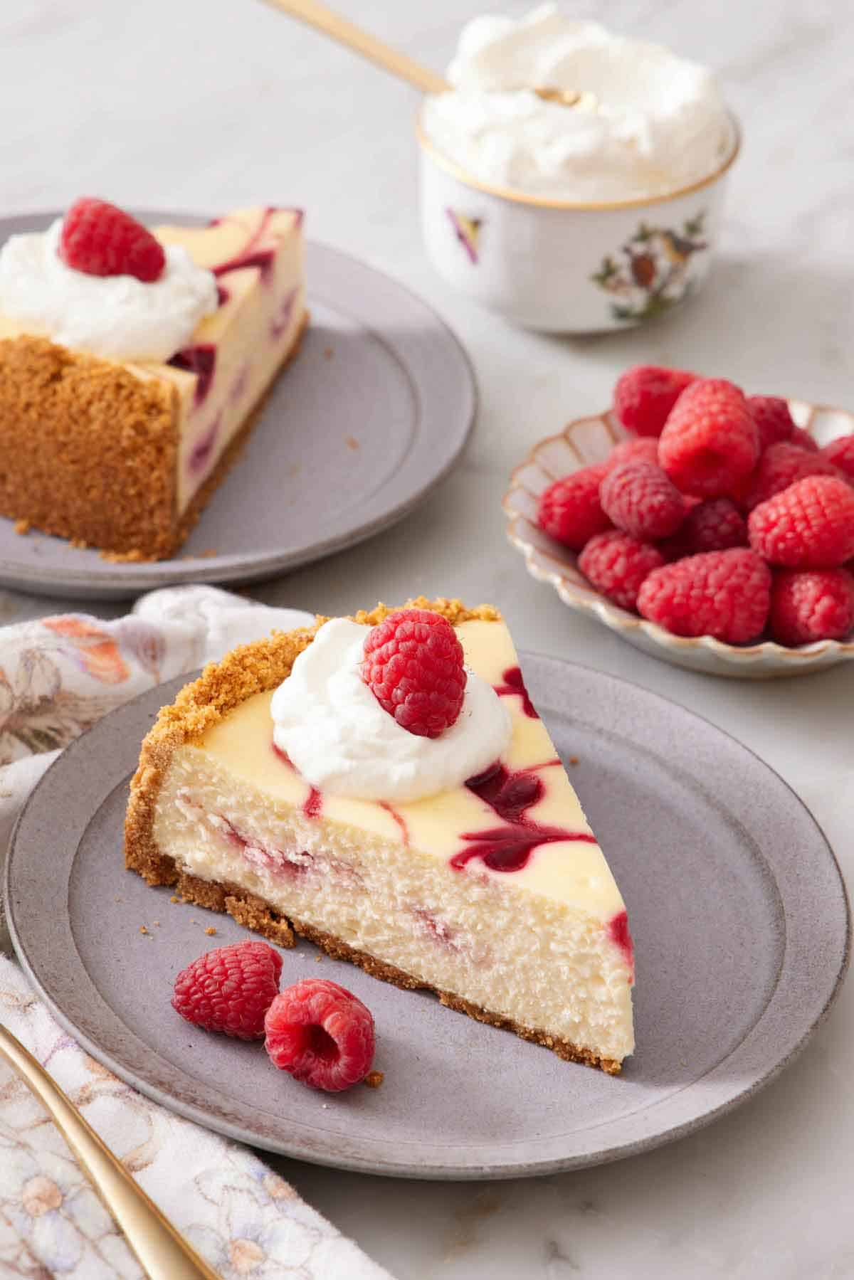 A slice of raspberry cheesecake topped with whipped cream and a raspberries. Another plates slice, a plate of raspberries, and a bowl of whipped cream in the background.