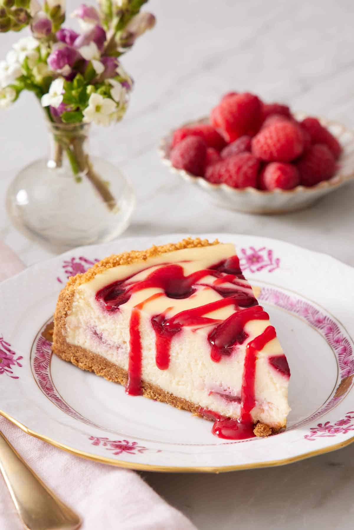 A slice of raspberry cheesecake on a plate with sauce drizzled on top. A vase of flowers and bowl of raspberries in the background.