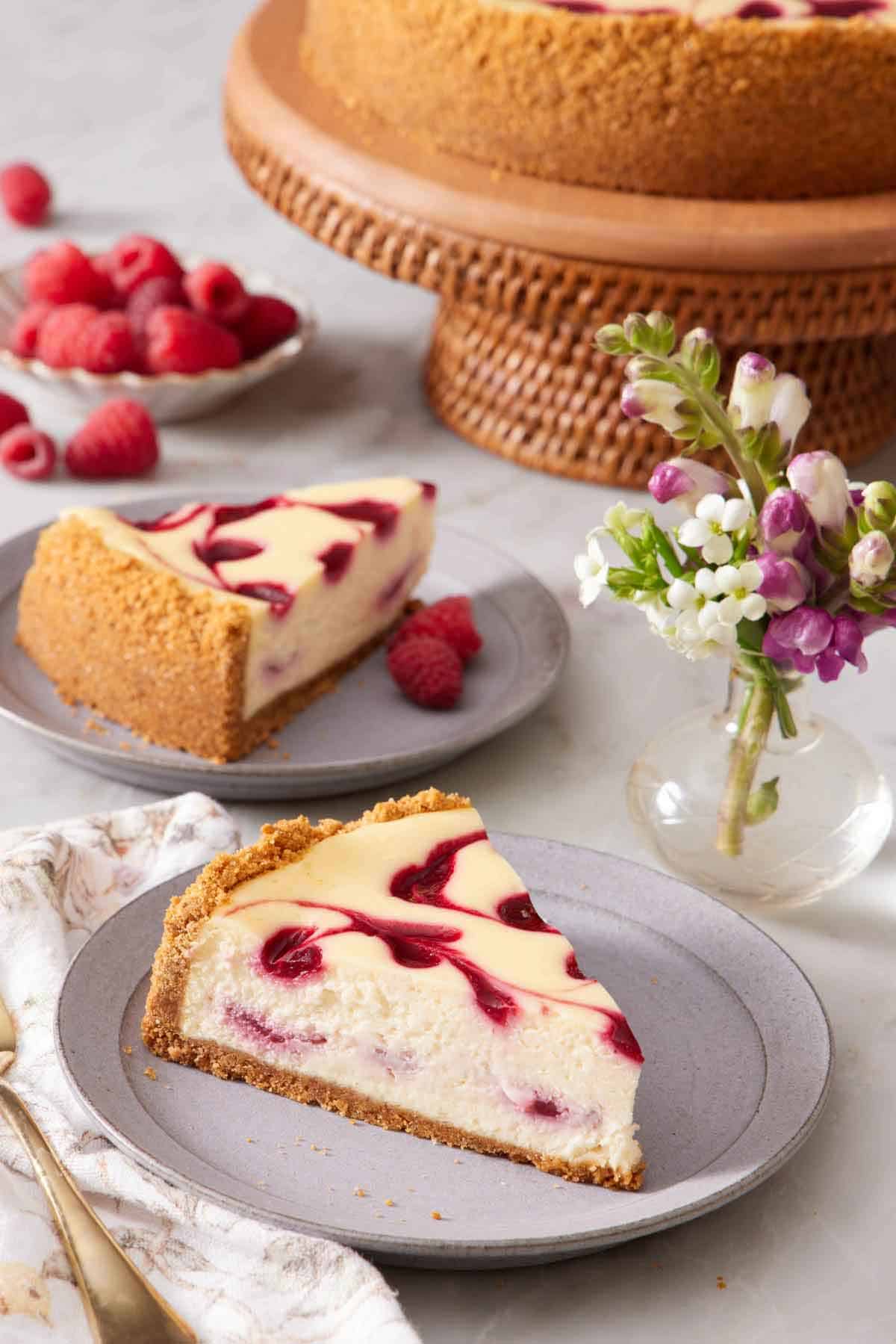 Two plates with slices of raspberry cheesecake. A mini vase of flowers with the rest of the cake in the background along with fresh raspberries.