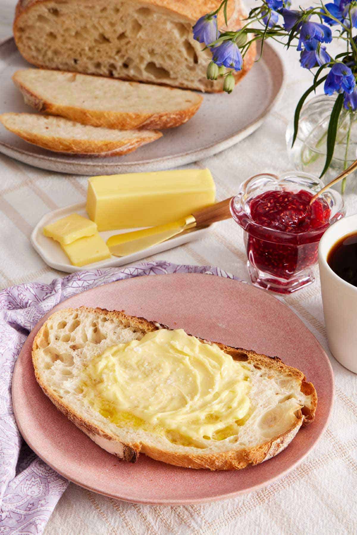 A plate with a slice of sourdough bread with butter spread on top. Jam, more butter, coffee, flowers, and a semi-cut loaf of sourdough bread in the back.