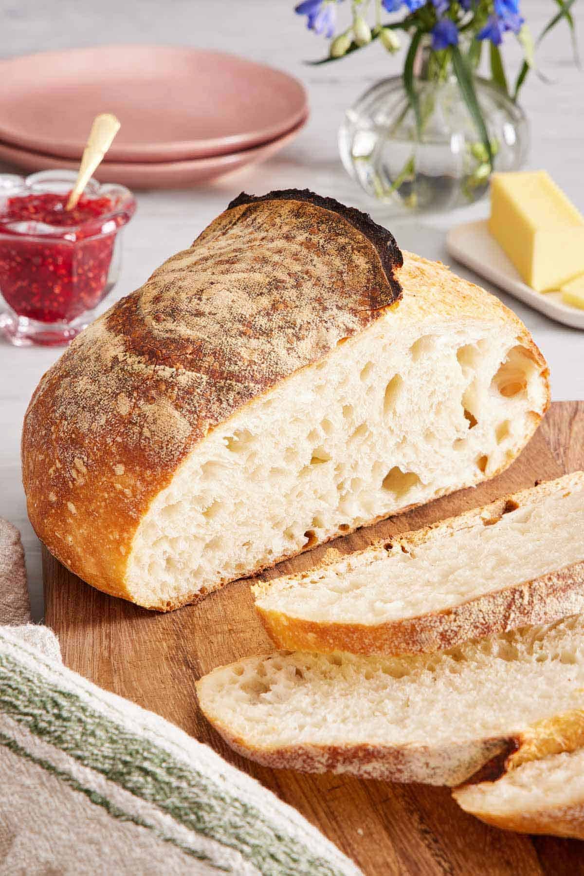 A loaf of sourdough bread on a wooden cutting board with a few slices cut in front. A jar of jam in the back with butter and plates.