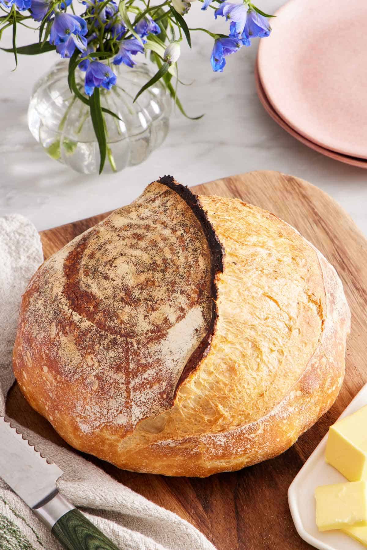 An uncut loaf of sourdough bread on a wooden serving board with some butter and flowers on the side.