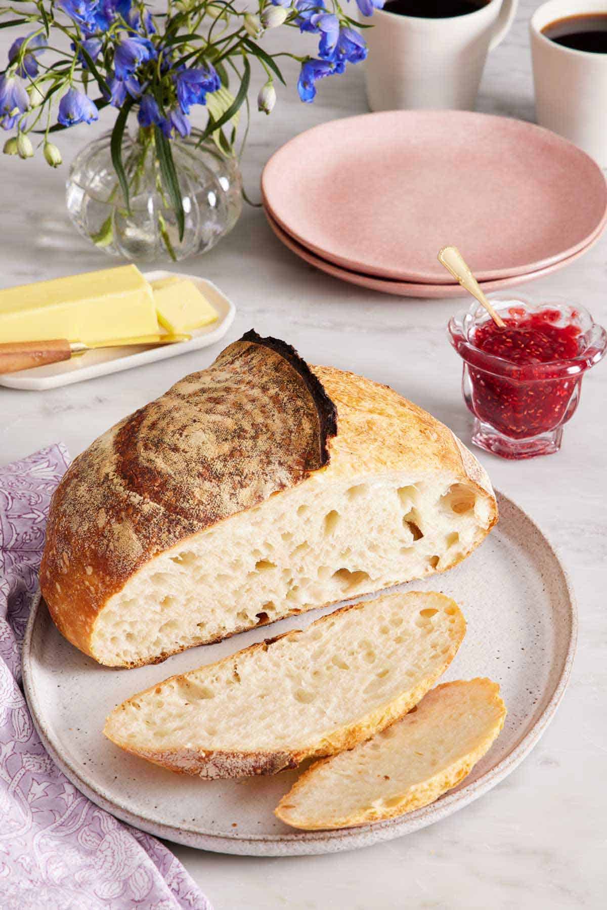 A plate with a loaf of sourdough bread with two slices cut off in front. A jar of jam, plates, butter, flowers, and two cups of coffee in the background.