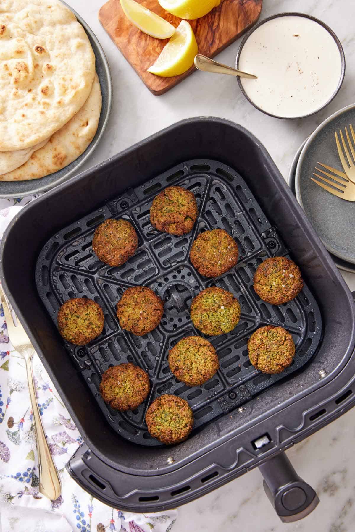 Overhead view of an air fryer basket with eleven air fryer falafel. Some pita bread in the background along with lemon wedges and a bowl of sauce.