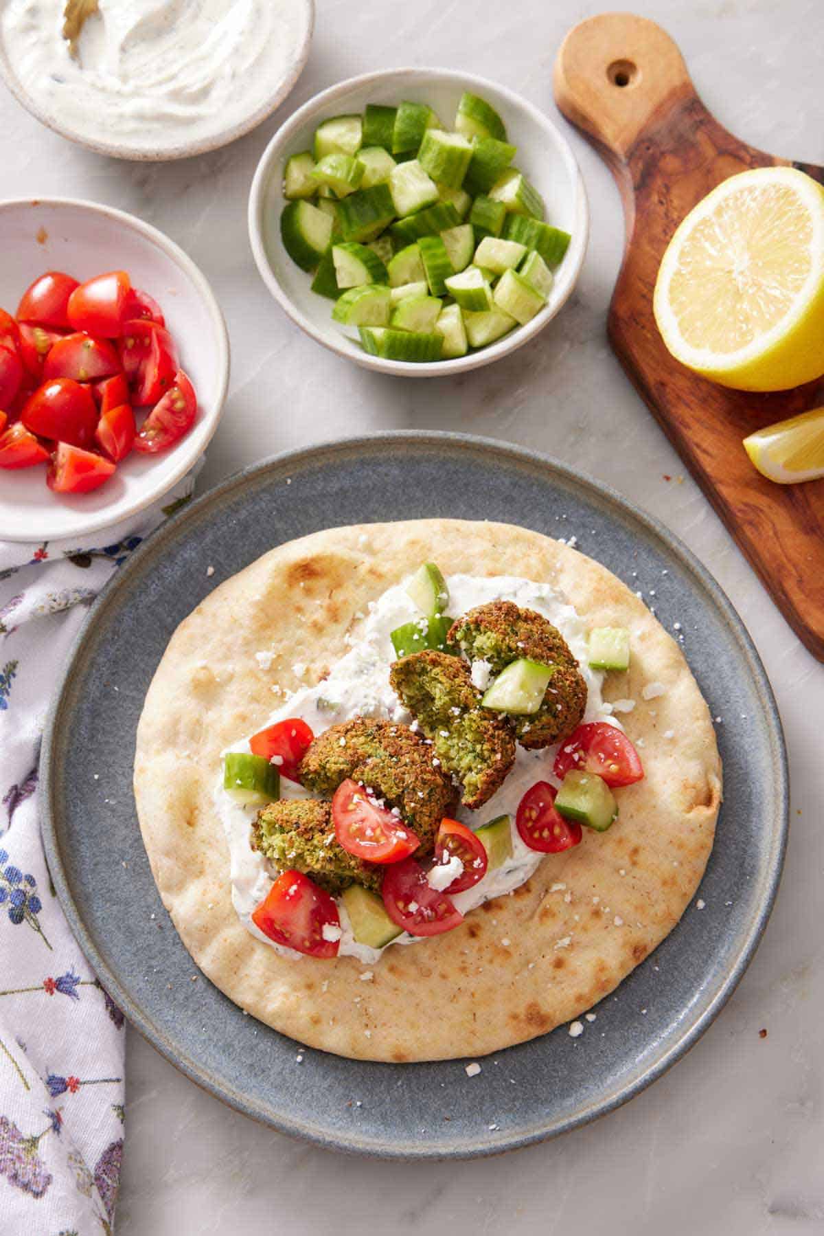 A plate with a pita bread with cut air fryer falafel with sauce, cut cucumbers, and tomatoes. Bowls of toppings and a cut lemon off to the side.