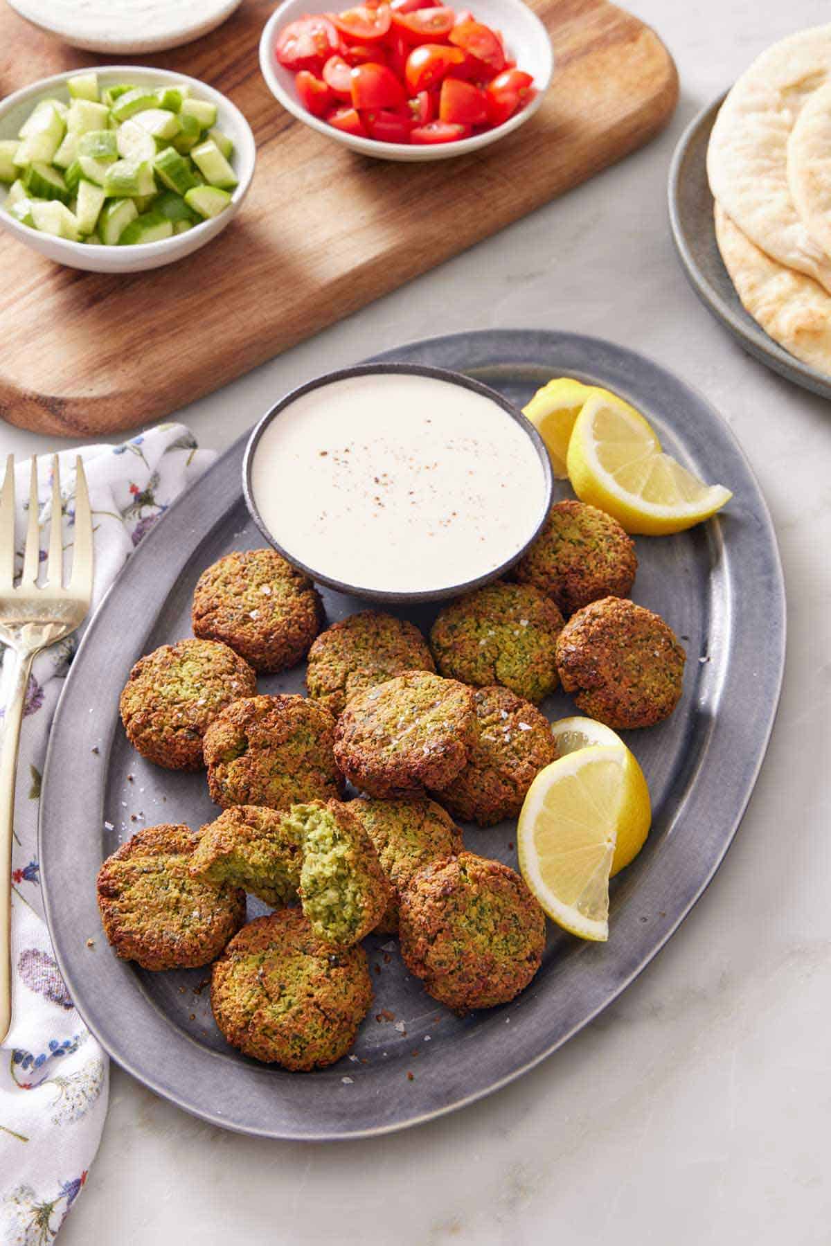 A platter of air fryer falafel with a bowl of dipping sauce and some lemon wedges. Pita bread in the background along with some cut tomatoes and cucumbers.