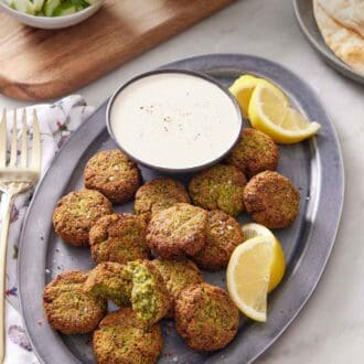 Pinterest graphic of a platter of air fryer falafel with a bowl of dipping sauce and some lemon wedges. Pita bread in the background along with some cut tomatoes and cucumbers.
