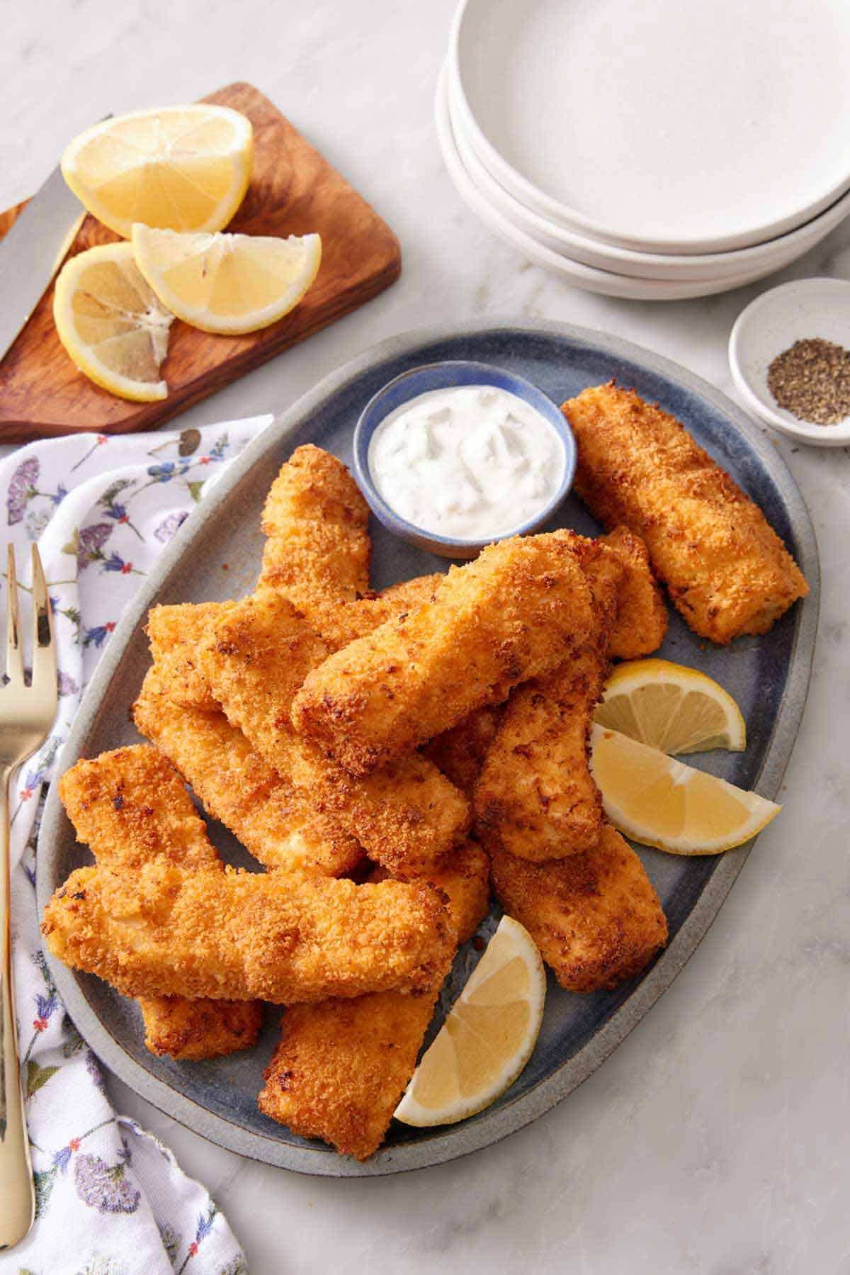 A platter of air fryer fish sticks with lemon wedges and a bowl of tartar sauce. A stack of plates. bowl of pepper, and cut lemon in the background.
