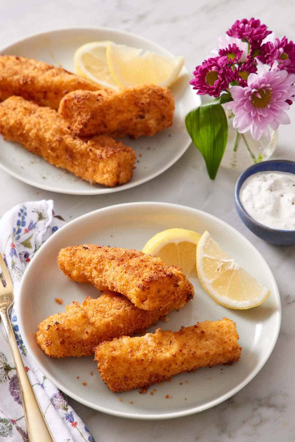 A plate with three air fryer fish sticks and lemon wedges. Another plate in the background along with a bowl of tartar sauce and vase of pink flowers.