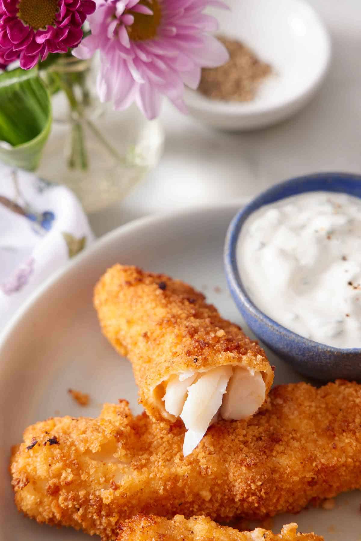 An air fryer fish stick cut opened, showing the flaky fish inside, propped up on another fish stick with a bowl of tartar sauce beside it.