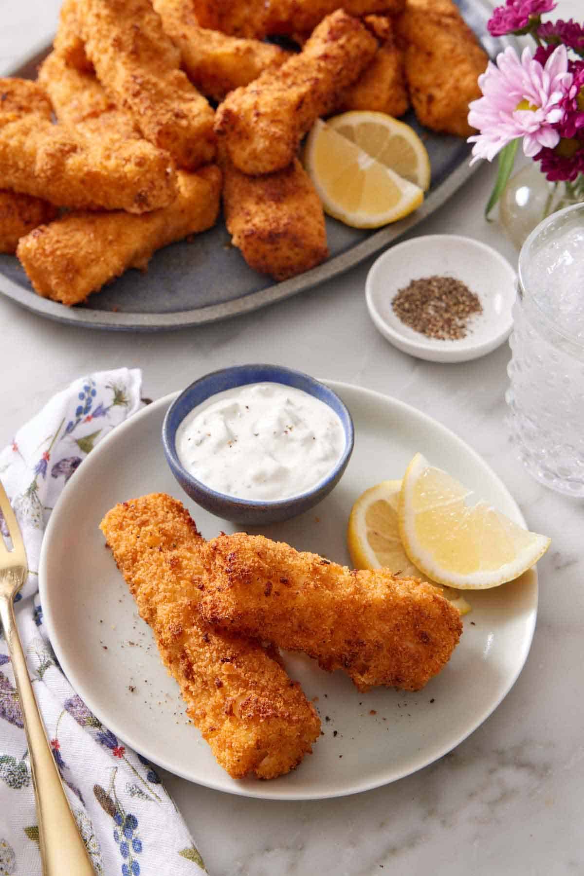 A plate with two air fryer fish sticks with lemon wedges and a bowl of tartar sauce. A platter of more air fryer fish sticks in the background along with a bowl of pepper and a drink.