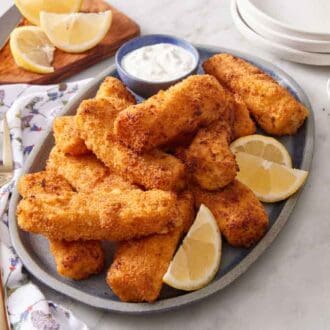 A pile of air fryer fish sticks with lemon wedges and bowl of tartar sauce on a platter. A fork, pepper, knife, and lemon wedges in the background.