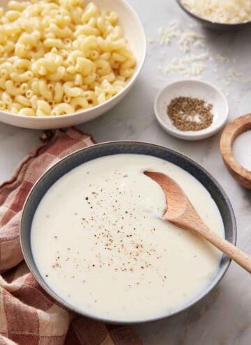 A bowl of béchamel sauce with a wooden spoon inside. Salt, Pepper, Cheese, and a bowl of macaroni in the background.