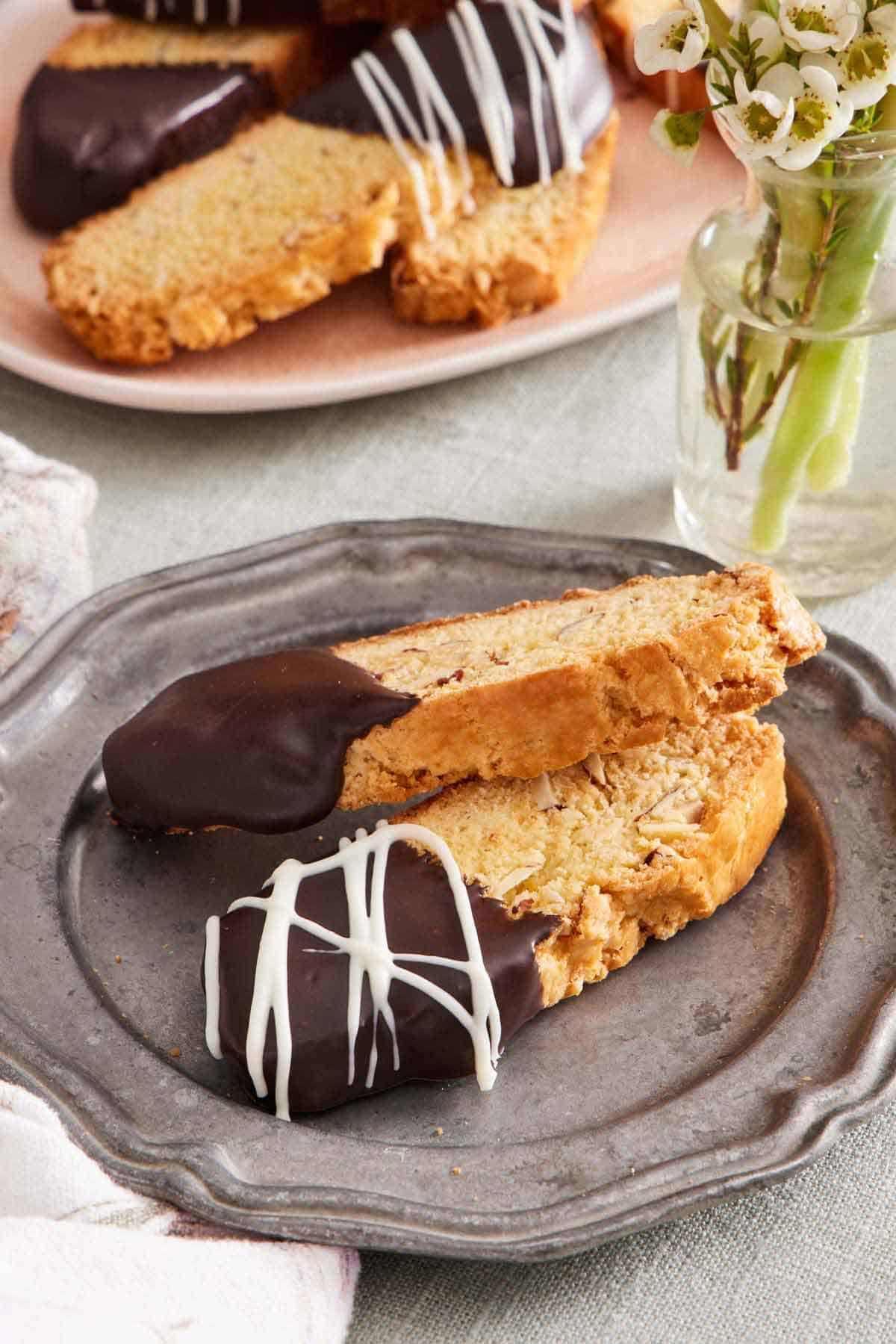 A plate with two biscotti, half dipped in chocolate. A vase of flowers and more biscotti on a platter in the background.