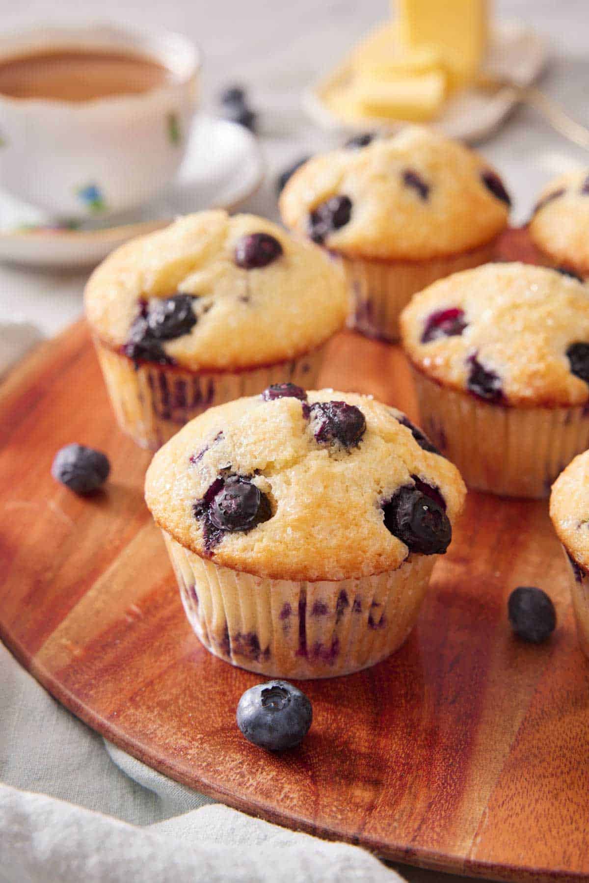 Multiple blueberry muffins on a wooden serving board. A cup of tea in the background along with butter.