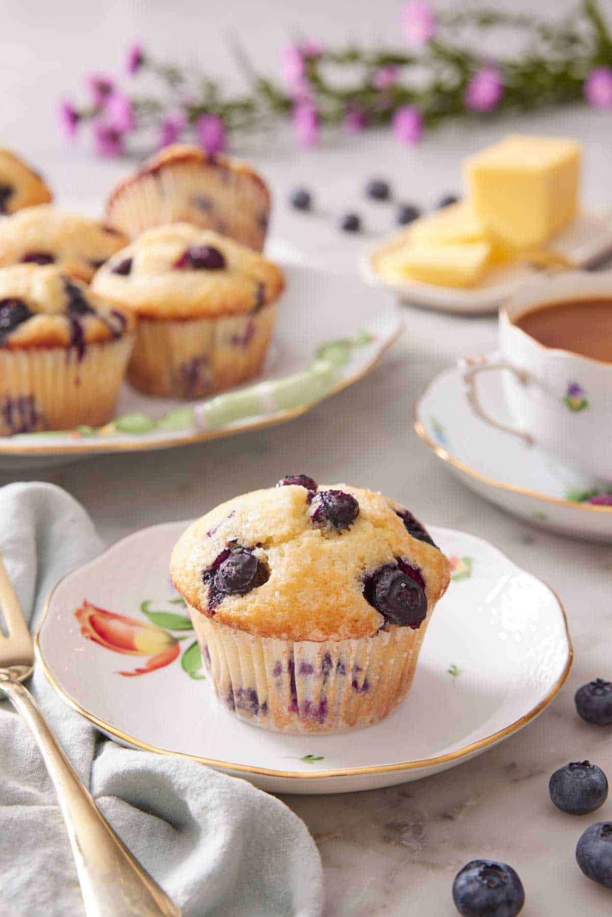 A plate with a single blueberry muffin. More muffins in the background on a platter with fresh blueberries scattered around with a cup of tea.