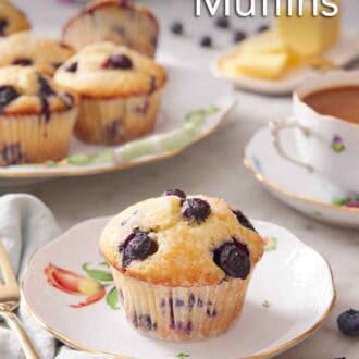 Pinterest graphic of a plate with a single blueberry muffin. More muffins in the background on a platter with fresh blueberries scattered around with a cup of tea.