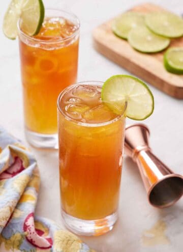 Two glasses of Dark and Stormy cocktails with a lime wheel on the edge. More lime wheels in the background along with a jigger.