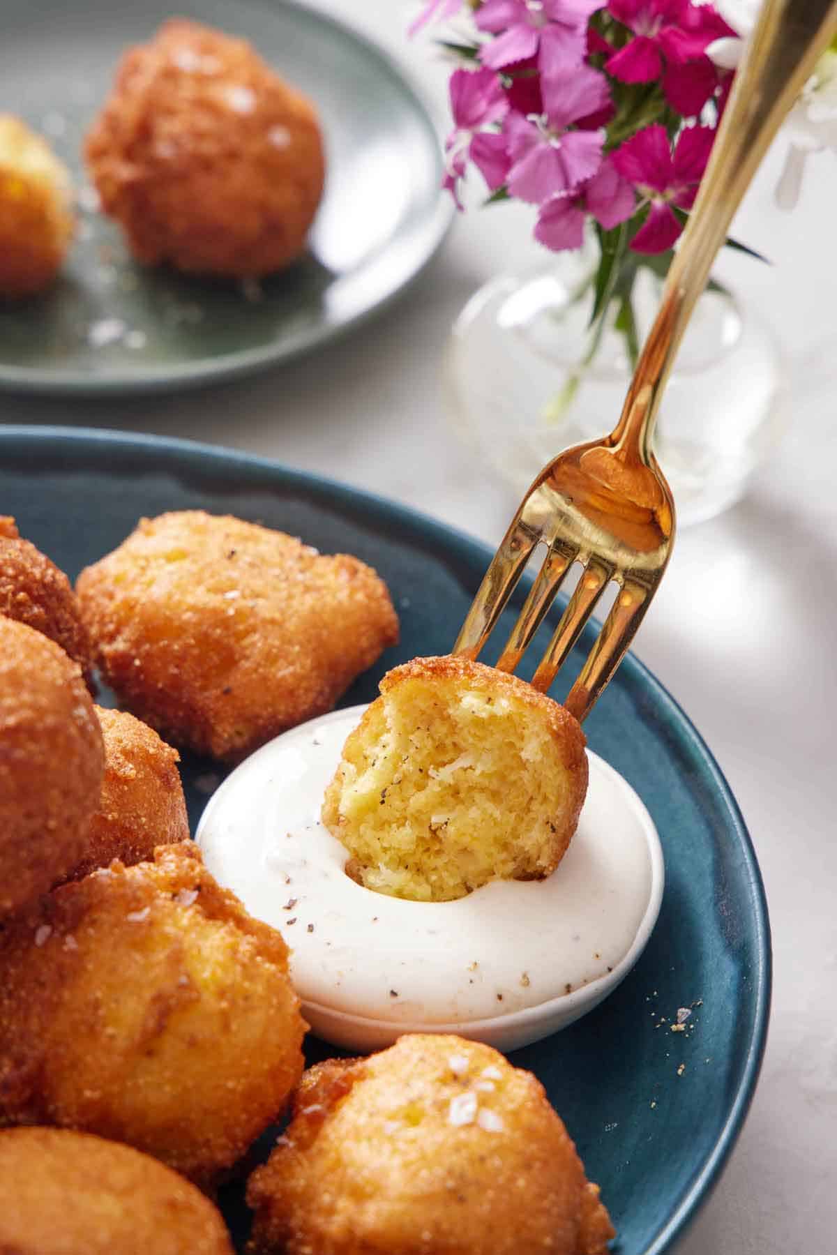 A plate of hush puppies with a fork dipping half of one into a small bowl of dipping sauce. Flowers in the background.