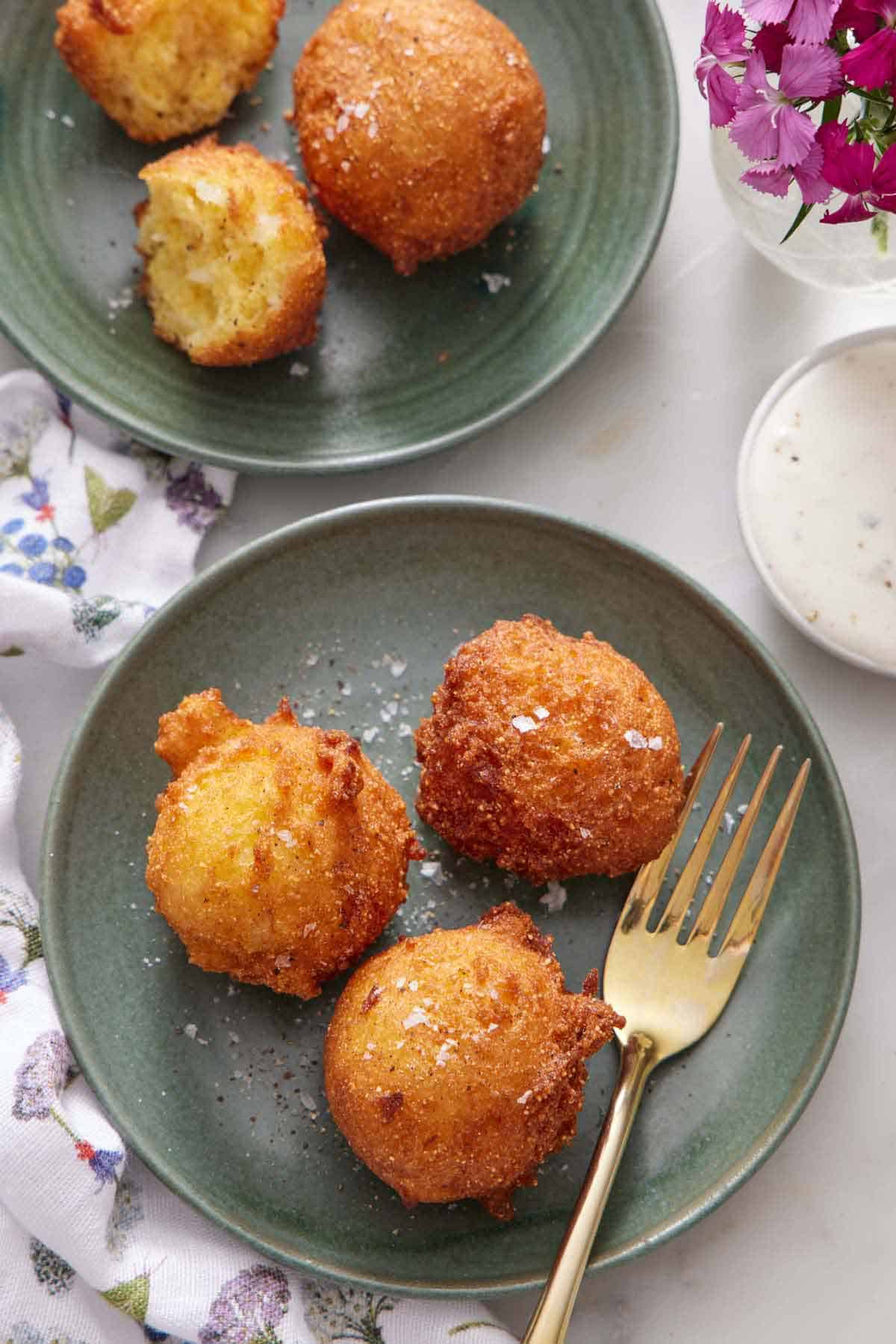 Overhead view of a plate with three hush puppies with a fork. Another plate with more hush puppies in the back.