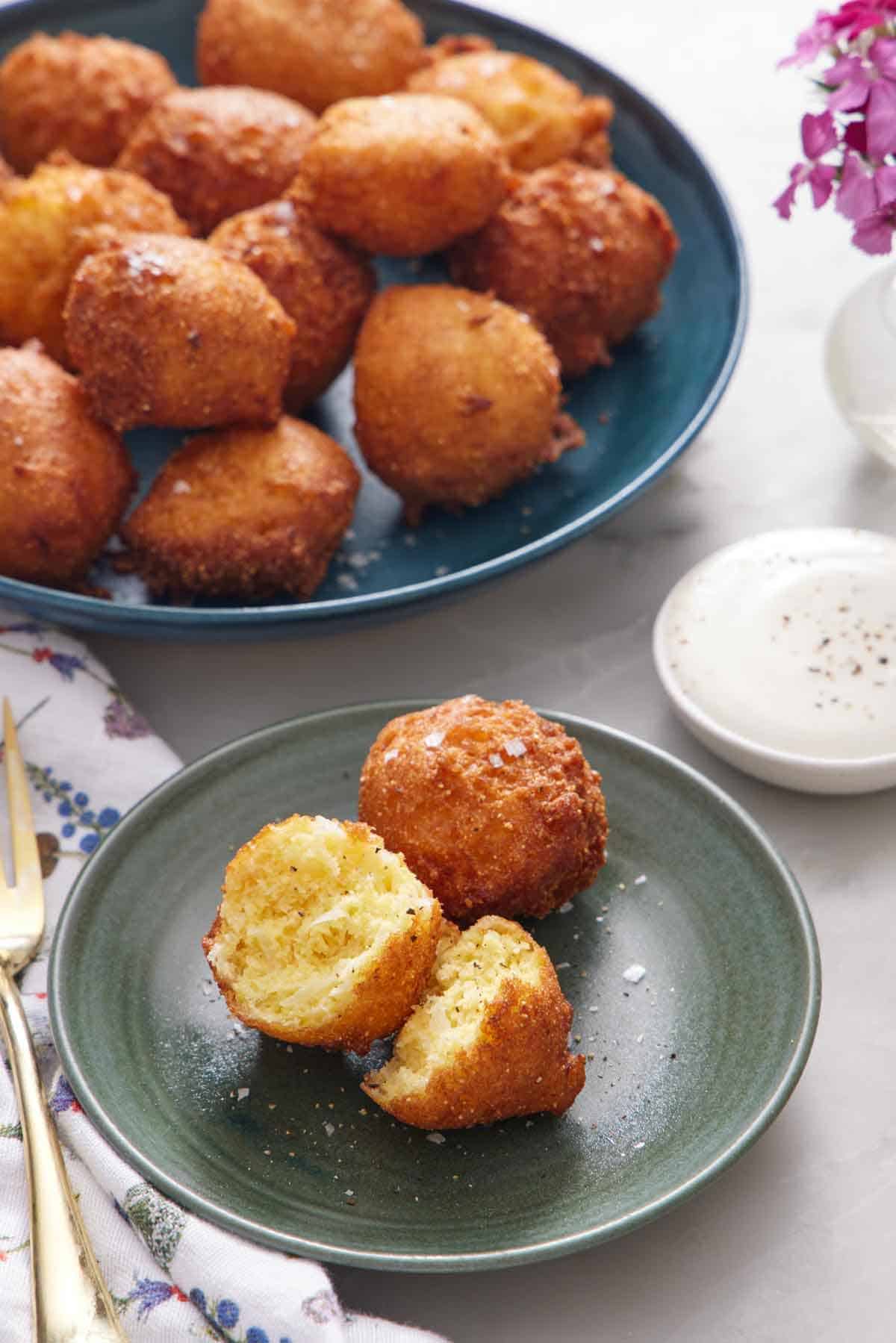 A green plate with two hush puppies, with one torn in half. A large plate of more hush puppies in the background along with a dipping sauce.