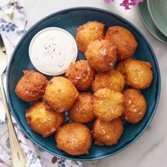 Overhead view of hush puppies with a small bowl of dipping sauce. A fork on a napkin on the side.